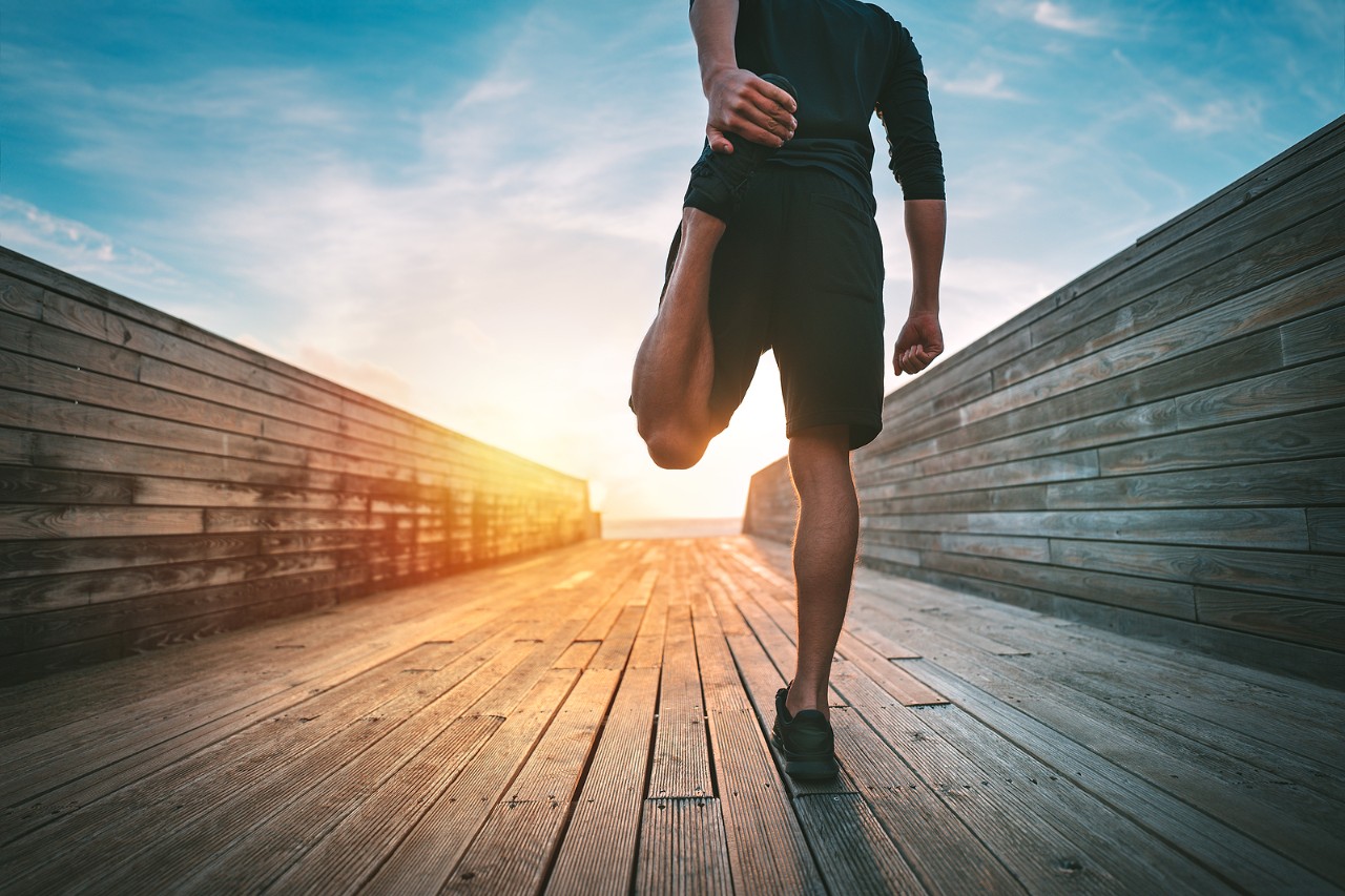 Young Slim Sporty Man Warming And Stretching Legs Before Run Outdoors At Sunset Or Sunrise. Athletic Young slim sporty man warming and stretching legs before run outdoors at sunset or sunrise. Athletic man in black sportswear doing fitness stretching exercises. Sport and healthy lifestyle.